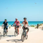 Vier personen fietsen met een helm op vlak voor een strand met heldere blauwe lucht in de achtergrond.