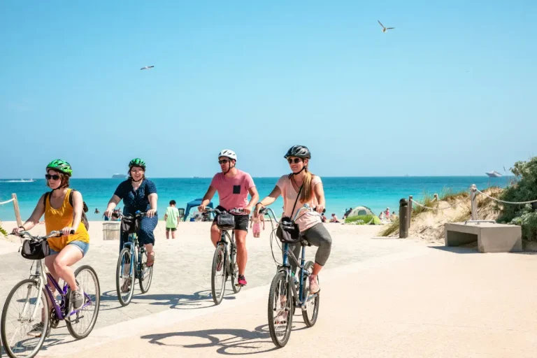 Vier personen fietsen met een helm op vlak voor een strand met heldere blauwe lucht in de achtergrond.