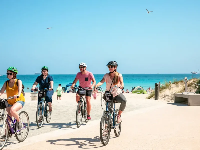 Vier personen fietsen met een helm op vlak voor een strand met heldere blauwe lucht in de achtergrond.