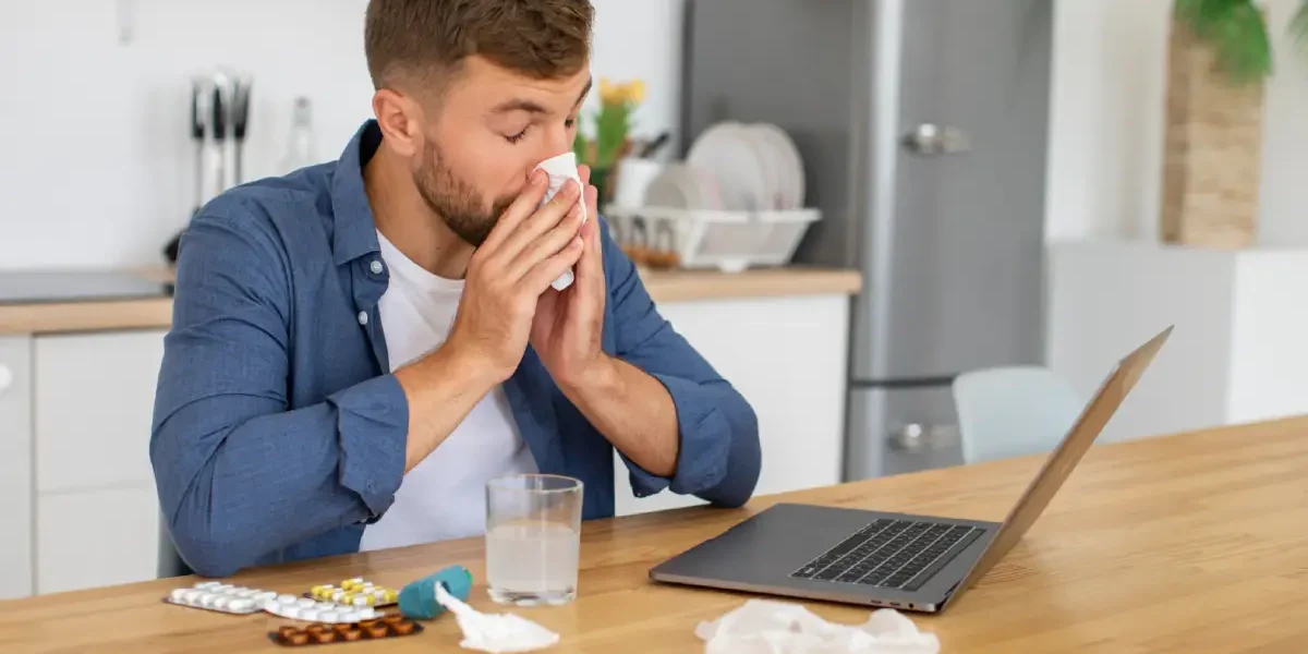 Een zieke man met verkoudheid is aan het werk op een laptop en snuit zijn neus.