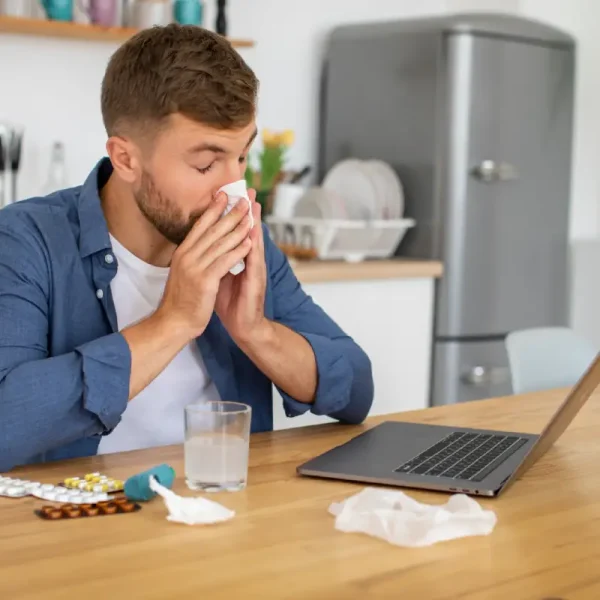 Een zieke man met verkoudheid is aan het werk op een laptop en snuit zijn neus.