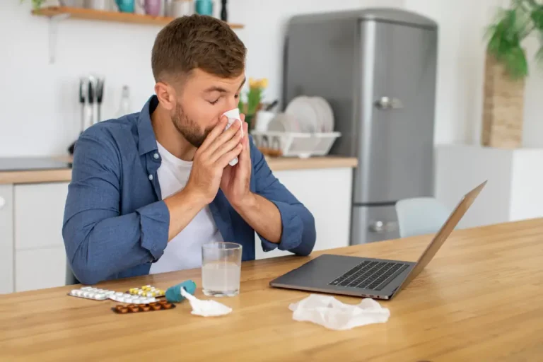 Een zieke man met verkoudheid is aan het werk op een laptop en snuit zijn neus.