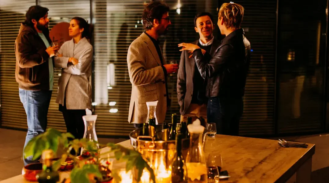 Mensen zijn aan het netwerken en staan rond een tafel met wijnflessen.